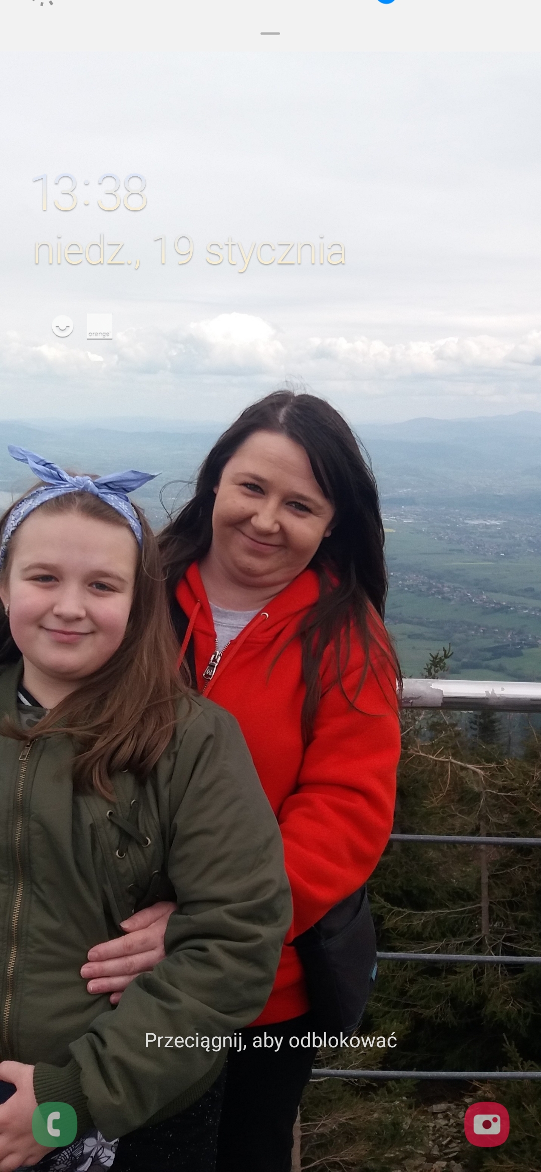 Dwie uśmiechnięte kobiety na tle górskiego krajobrazu, widoczne z platformy widokowej. Jedna w czerwonej bluzie, druga w zielonej kurtce z niebieską opaską.