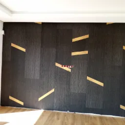 Ciemna ściana wykończona panelami z drewnopodobną fakturą i złotymi, geometrycznymi wstawkami, widoczne gniazdka elektryczne, fragment podłogi i drzwi.