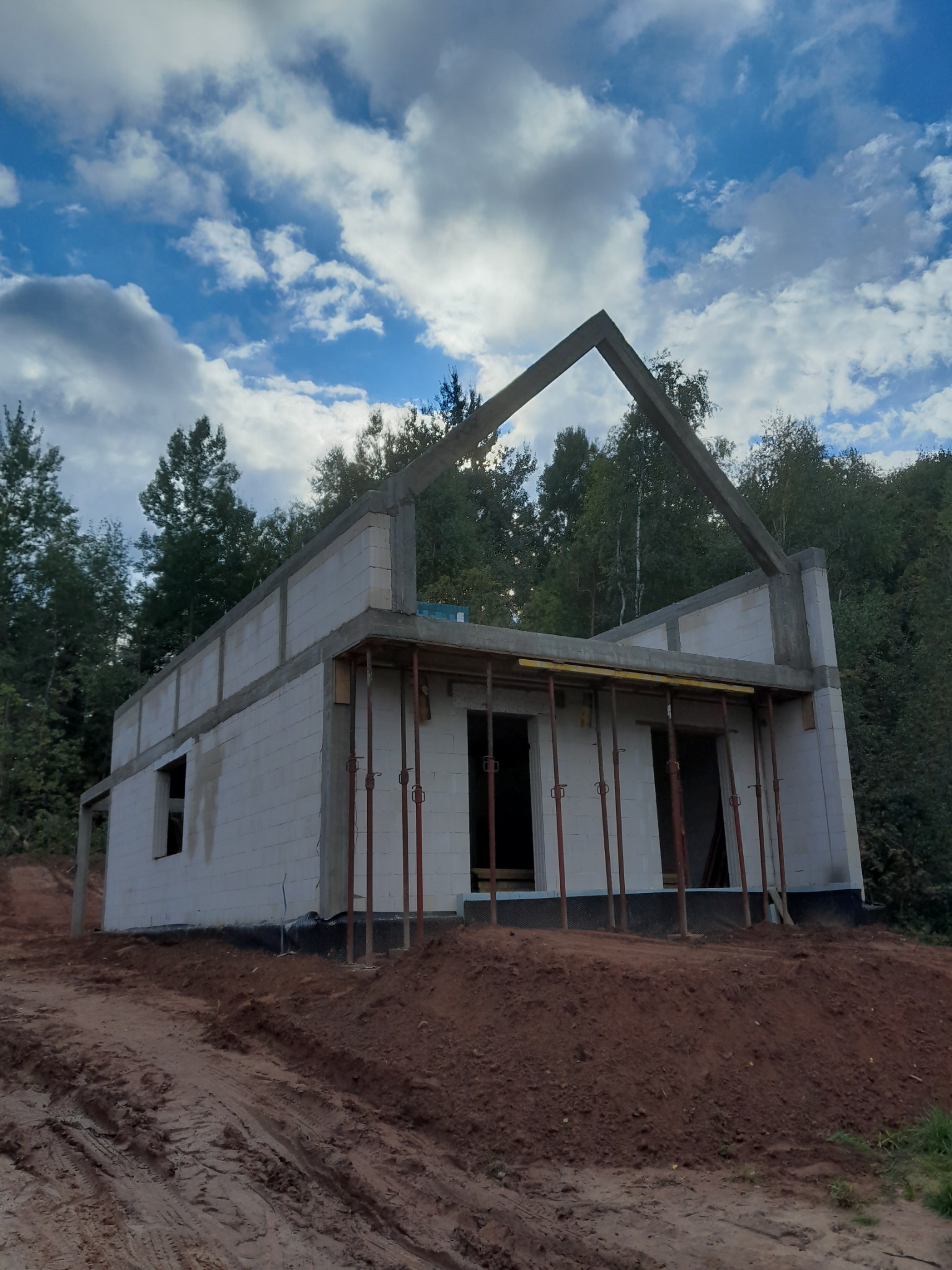 Surowy stan domu z betonu komórkowego w trakcie budowy, widoczne podpory stropu, grunt wokół budynku, las w tle i pochmurne niebo.
