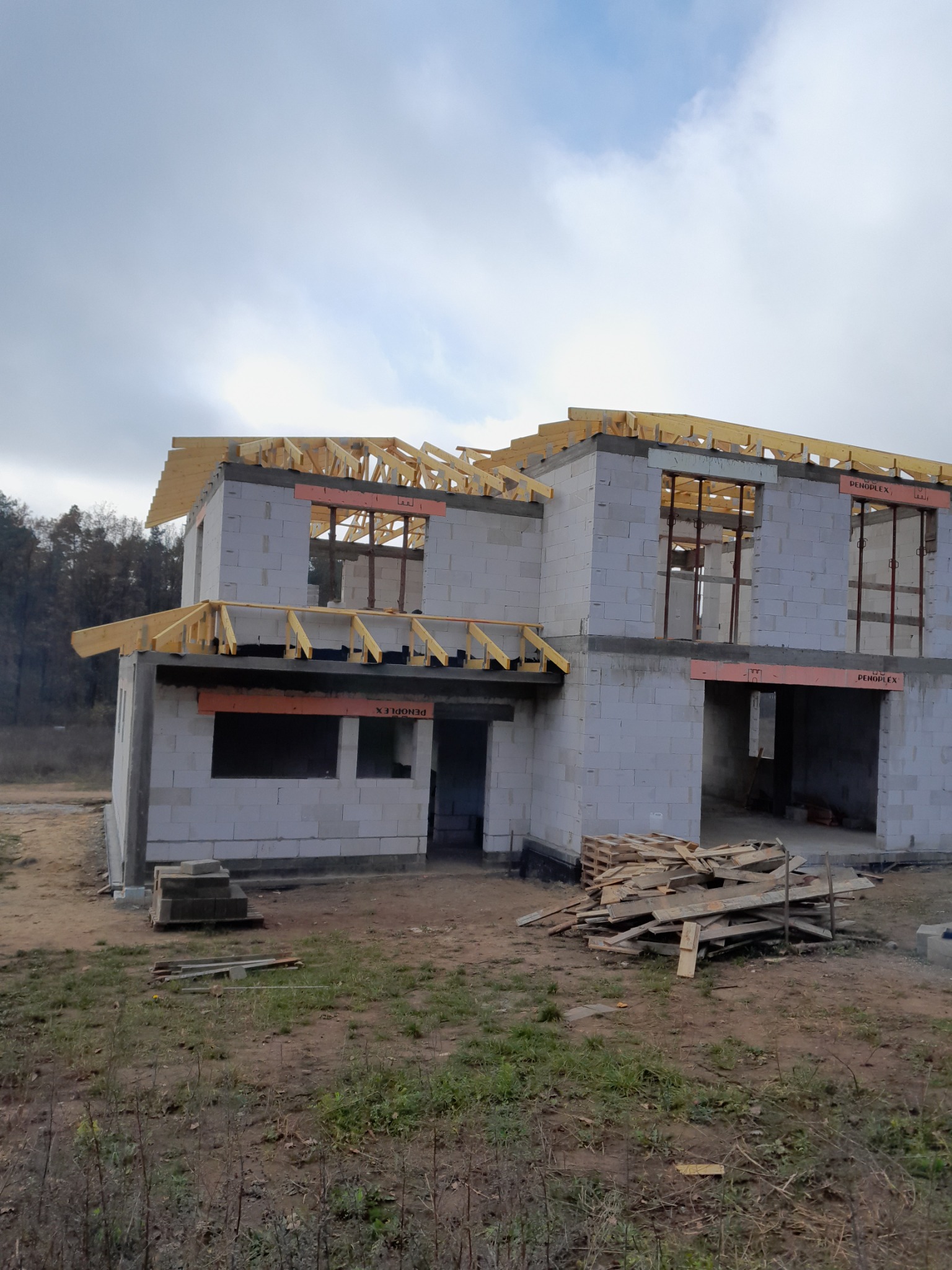 Surowy stan dwukondygnacyjnego domu z pustaków, z widoczną konstrukcją dachu z belek, częściowo zabezpieczoną pomarańczową pianką, z paletą i stertą desek na pierwszym planie.