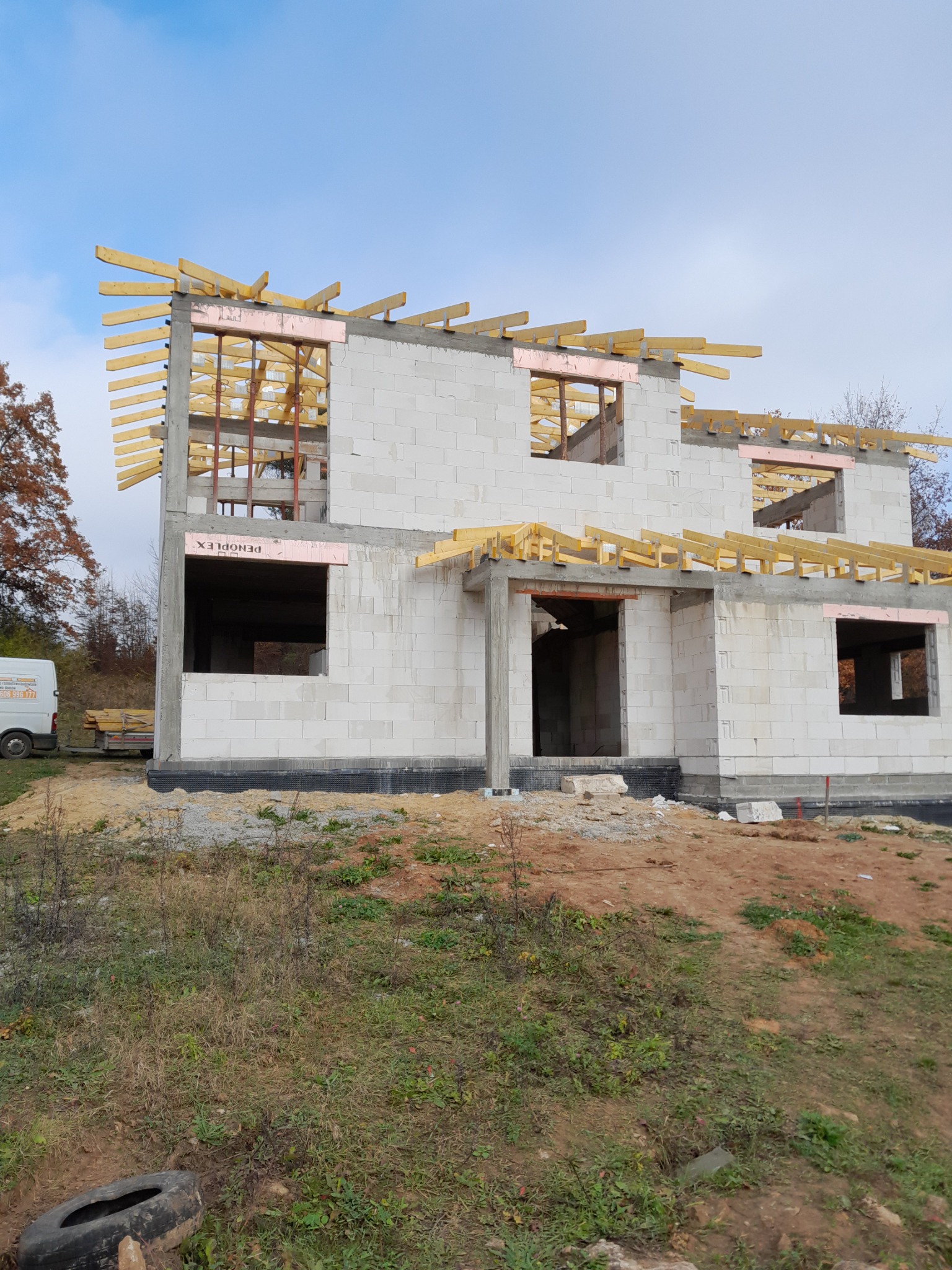 Wznoszenie dwukondygnacyjnego budynku z białych bloczków, widoczne drewniane elementy konstrukcyjne dachu i otwory okienne zabezpieczone różową pianką, na pierwszym planie ziemia i trawa, w tle...