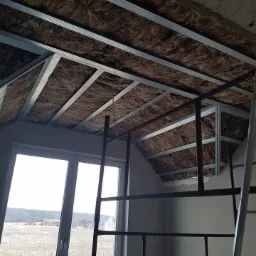 Montaż izolacji z wełny mineralnej w metalowym stelażu sufitu poddasza, widok z rusztowania, okno w tle.