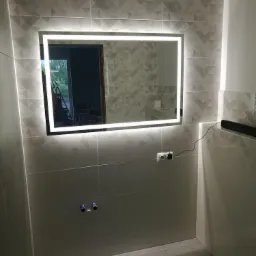 Wykończona łazienka z podświetlanym lustrem na ścianie z geometrycznymi płytkami, widoczne przyłącza wodne i gniazdko elektryczne z podłączonym kablem.