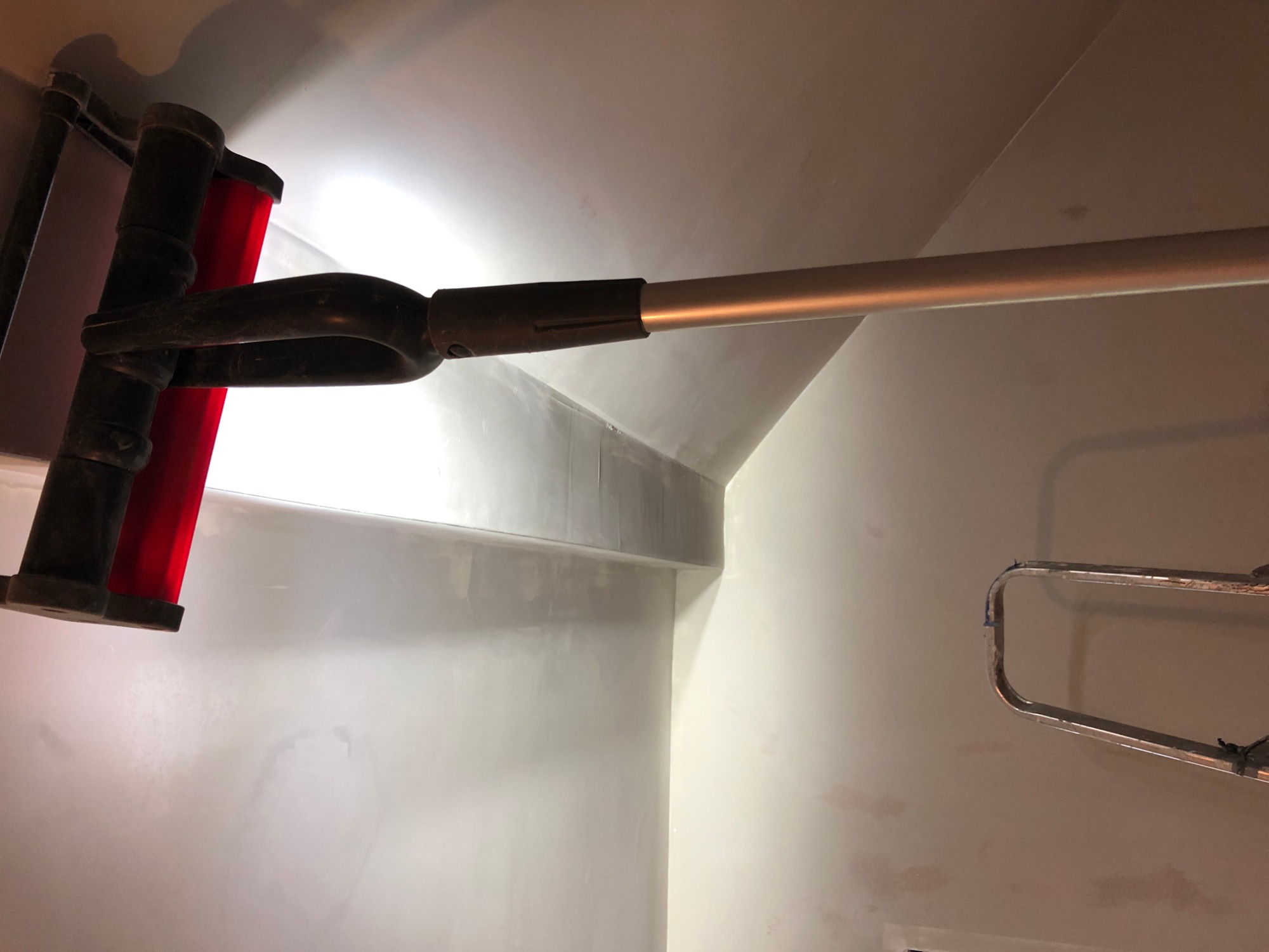 Wałek z czerwoną nakładką na teleskopowym kiju podczas nakładania gładzi na ścianę i sufit w rogu pomieszczenia, widoczny fragment drabiny.