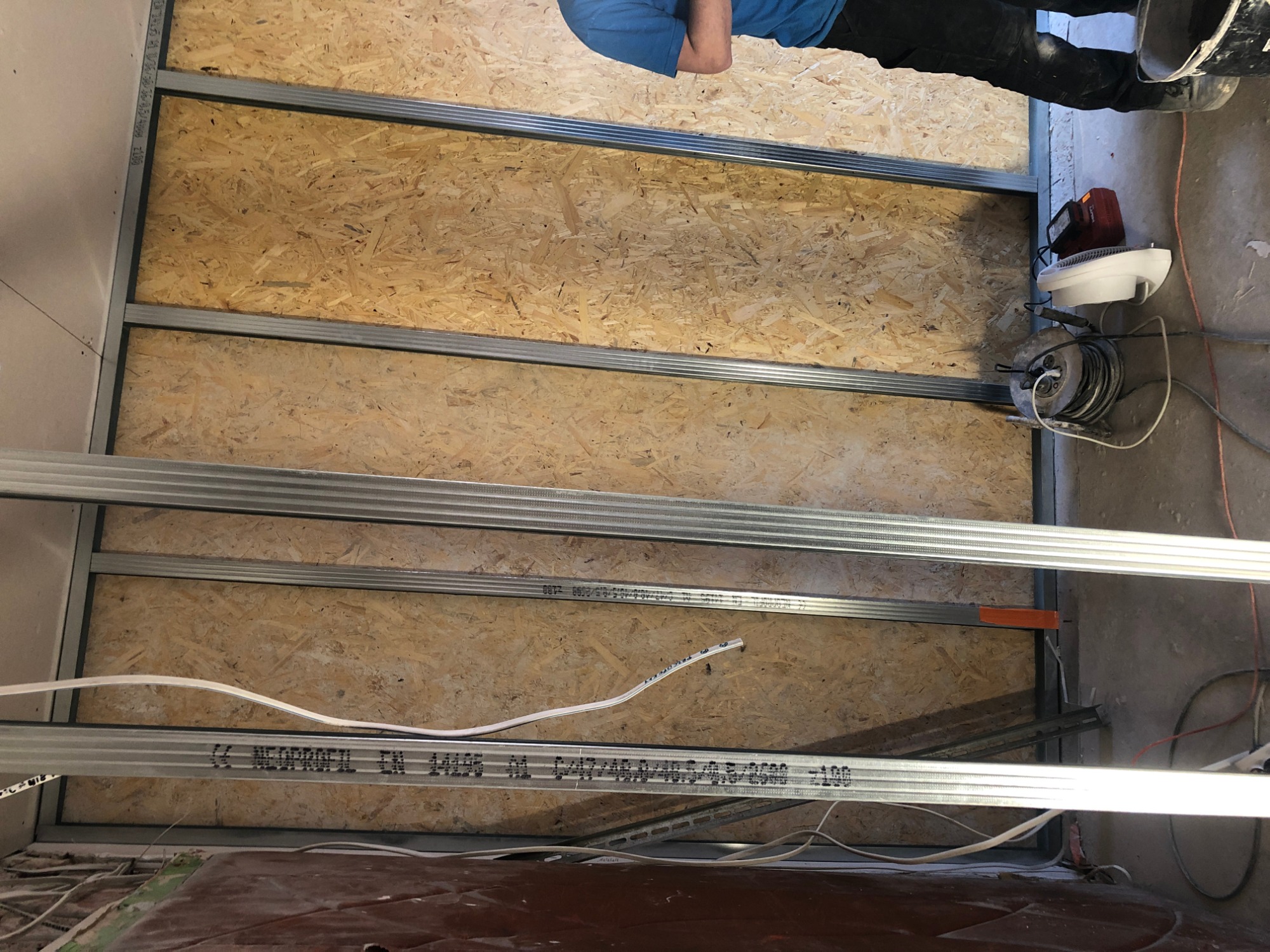 Konstrukcja ściany działowej z płyty kartonowo-gipsowej, widoczne profile metalowe, płyta OSB, instalacja elektryczna wewnątrz, narzędzia i fragment osoby w tle.