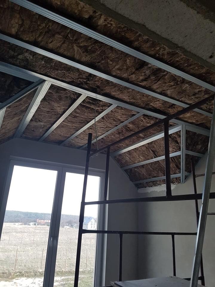 Montaż izolacji z wełny mineralnej w metalowym stelażu sufitu poddasza, widok z rusztowania, okno w tle.