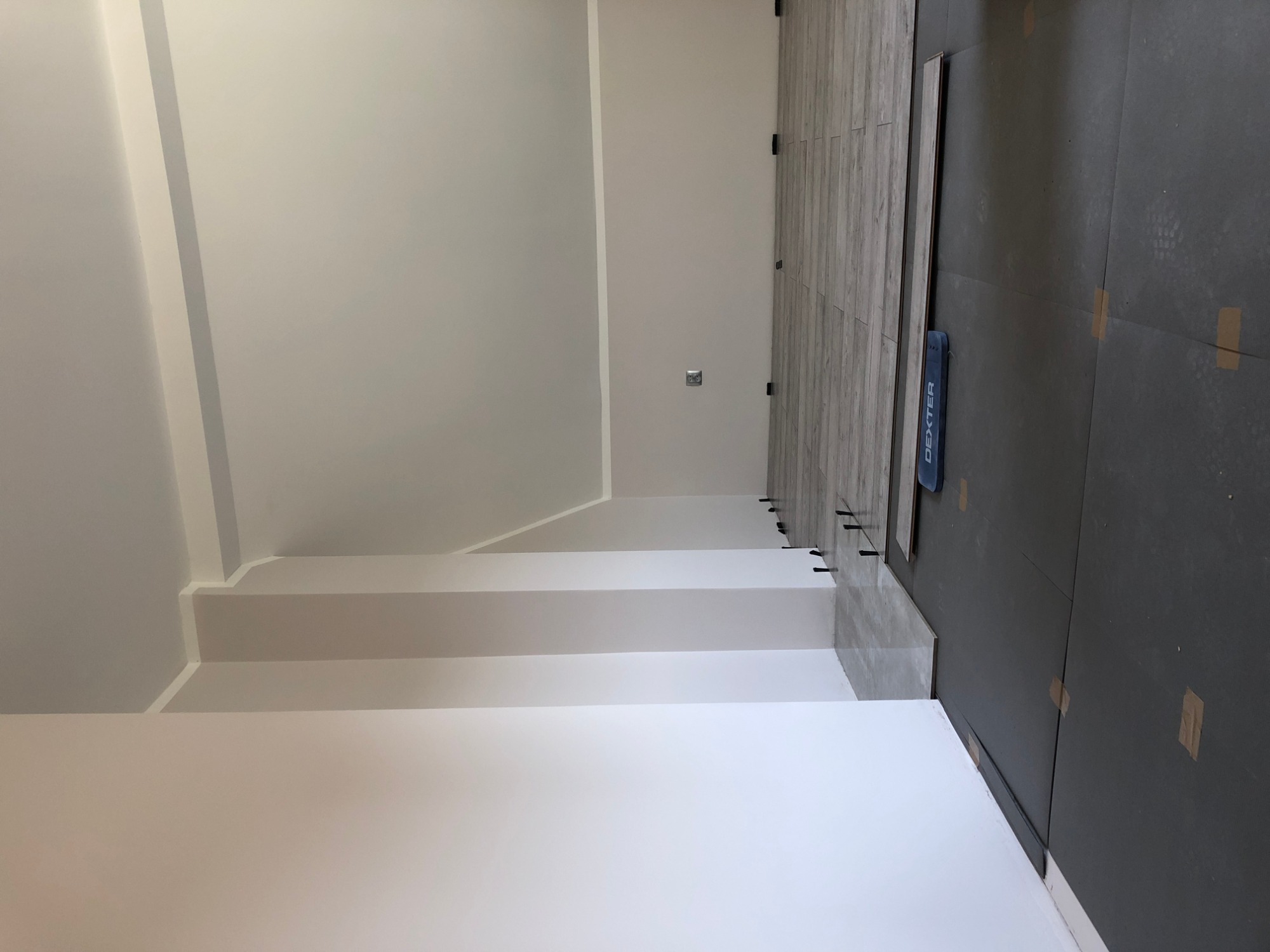 Wnętrze w trakcie prac wykończeniowych: świeżo pomalowana biała ściana z widocznymi schodkowymi elementami, częściowo ułożona podłoga z paneli imitujących drewno oraz czarna mata pod panele...