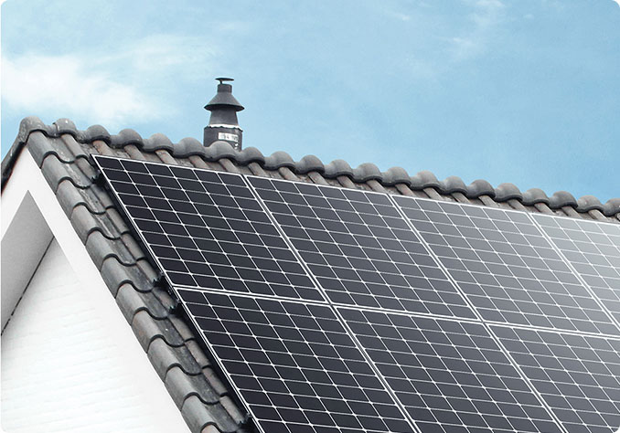 Panel słoneczny zamontowany na dachu pokrytym starą dachówką, z widocznym kominem w tle i błękitnym niebem.