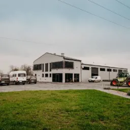 Nowoczesny budynek firmy z zaparkowanymi samochodami dostawczymi i traktorem, na tle zielonego trawnika w pochmurny dzień.