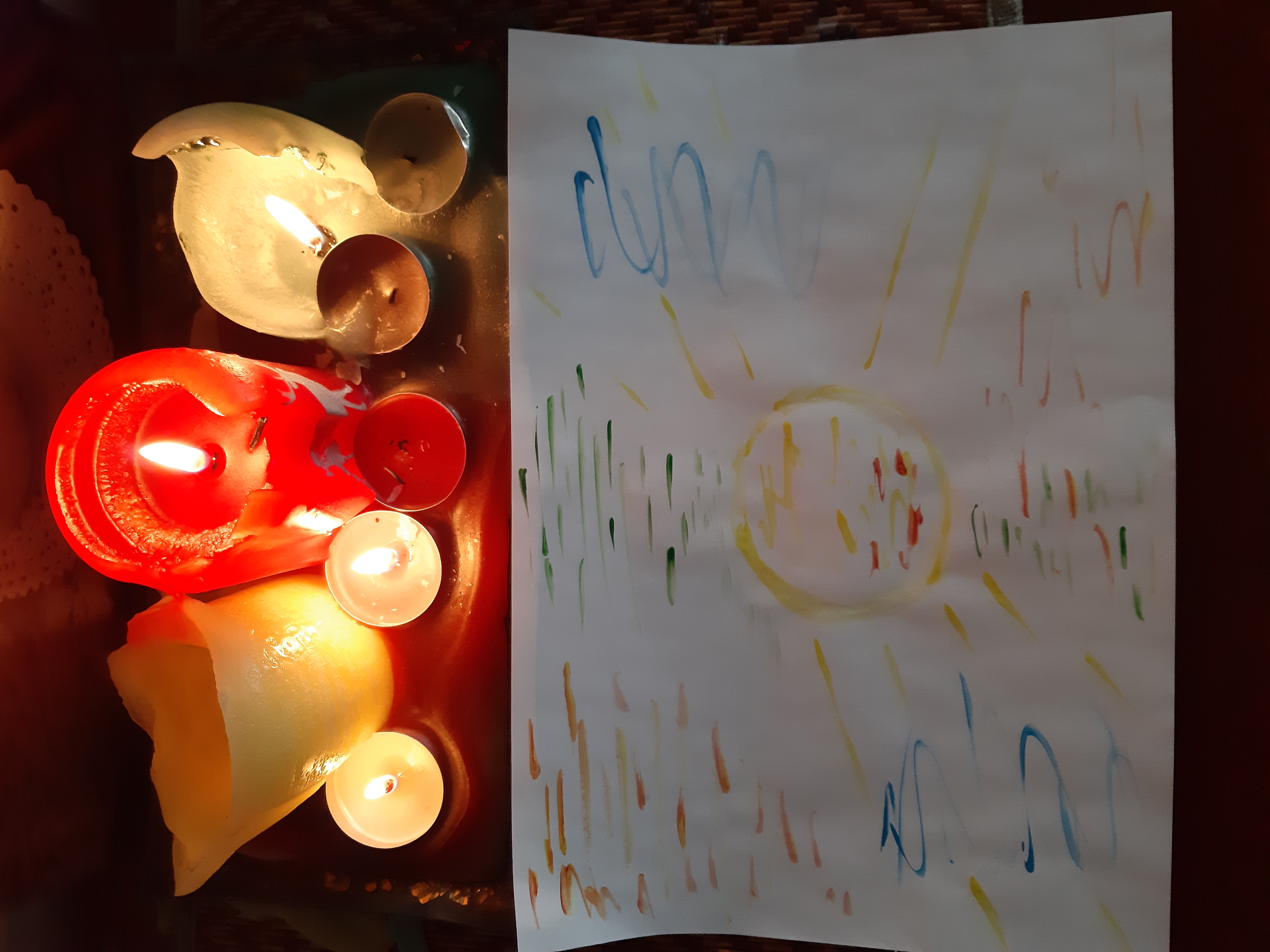 Aromatyczne świece w różnych kolorach i kształtach, obok dziecięcy rysunek słońca z promieniami, tworzą relaksującą atmosferę.