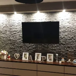 Ściana w salonie wykończona szarą kamienną okładziną, podświetlona punktowymi lampami sufitowymi, z zawieszonym telewizorem i komodą z dekoracjami świątecznymi i ramkami na zdjęcia.