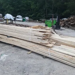 Stos desek z jasnego drewna kominkowego związany białym sznurkiem, na tle placu składowego z wózkiem widłowym i stertą drewna opałowego.