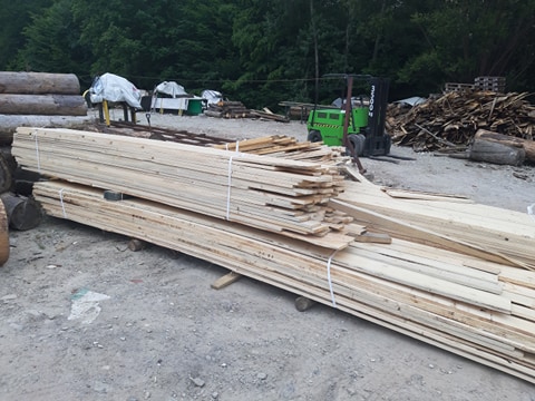 Stos desek z jasnego drewna kominkowego związany białym sznurkiem, na tle placu składowego z wózkiem widłowym i stertą drewna opałowego.