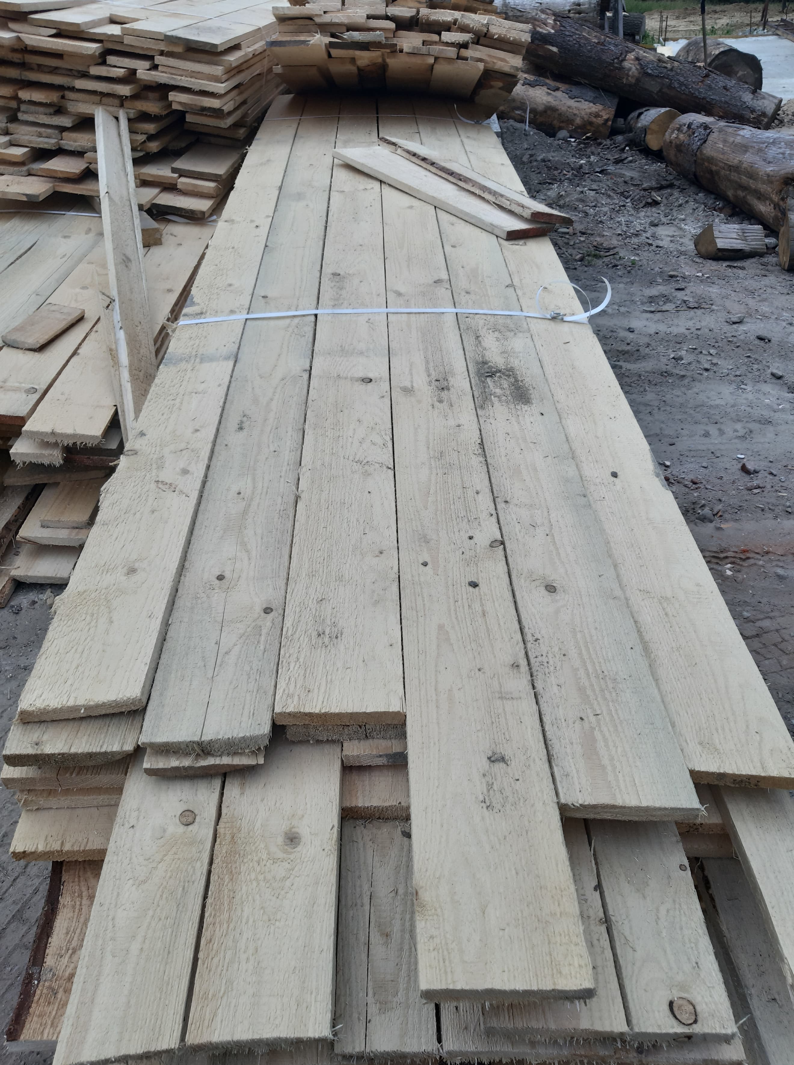 Stos jasnych, surowych desek ułożonych na ziemi, związanych białą taśmą, z widocznymi sękami i naturalnym usłojeniem drewna.