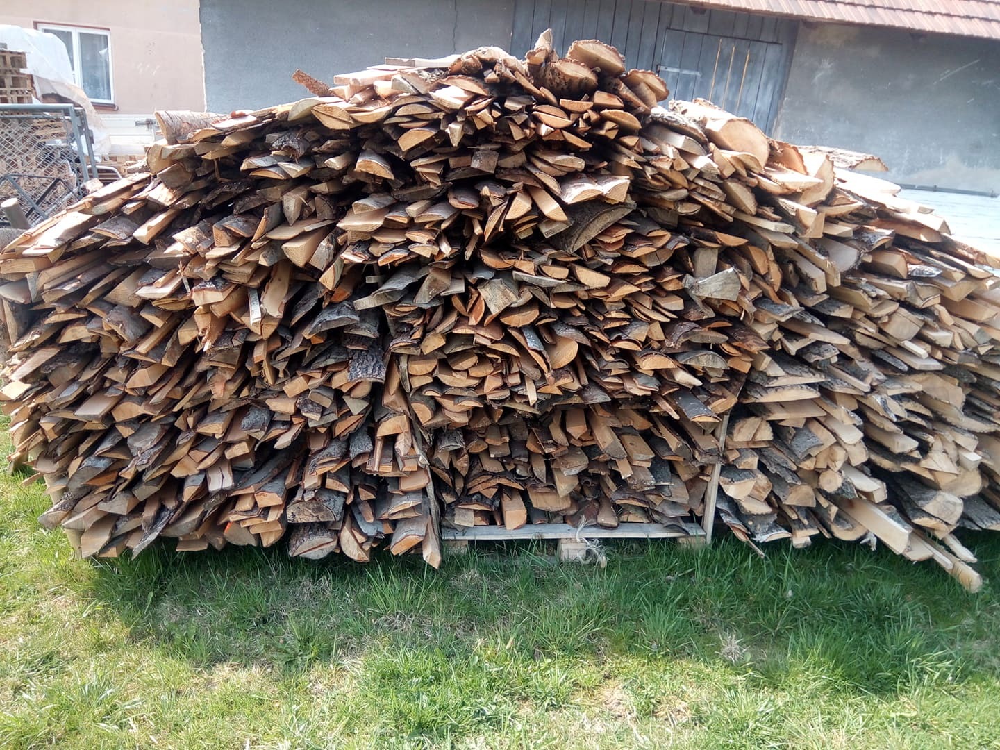Stos pociętego drewna na palecie, ułożonego na trawie przed budynkiem gospodarczym.
