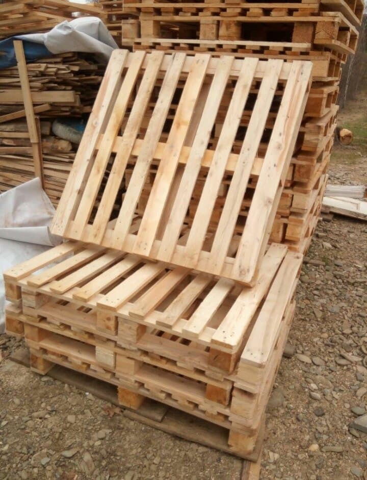 Stos jasnych, drewnianych palet ułożonych na żwirowym podłożu, z luźno ułożoną paletą na wierzchu i stosem drewna w tle.