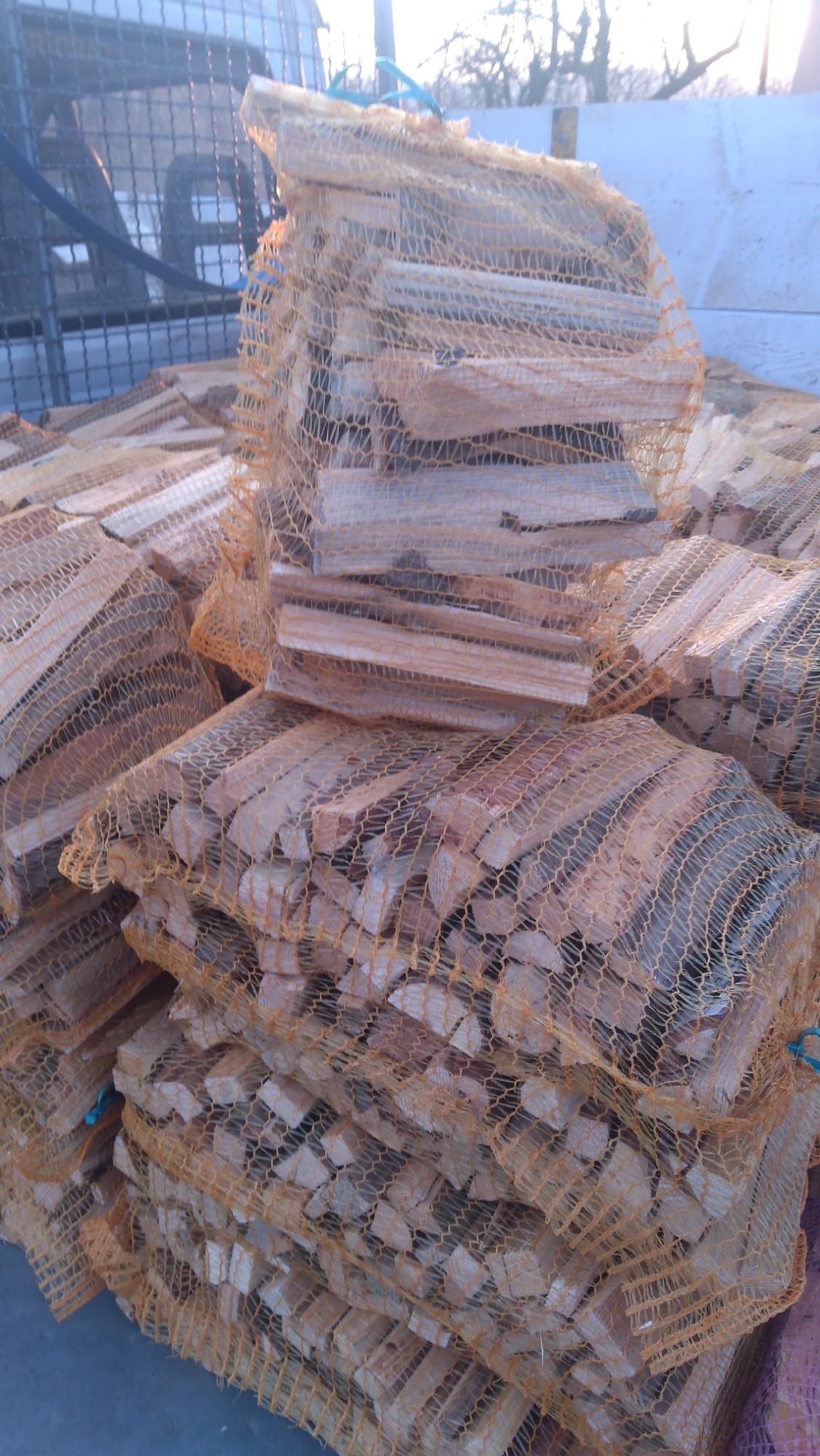 Stos worków z pociętym drewnem opałowym, zabezpieczonych pomarańczową siatką, ułożonych na pace samochodu.