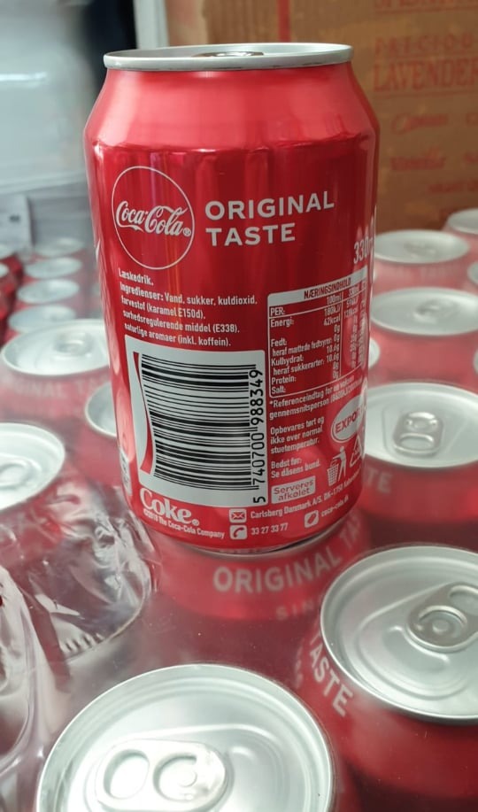 Puszka Coca-Coli Original Taste z kodem kreskowym i informacjami o wartościach odżywczych, otoczona innymi puszkami, z widocznym elementem opakowania zbiorczego.
