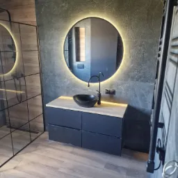 Nowoczesna łazienka z kabiną prysznicową typu walk-in, okrągłym lustrem z podświetleniem LED, czarną umywalką nablatową i szafką z drewnianym blatem na tle ściany z szarego kamienia.