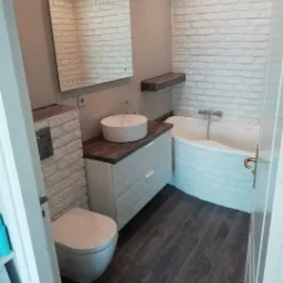 Nowoczesna łazienka z białymi płytkami imitującymi cegłę, umywalką nablatową na szafce z frezowanymi frontami, lustrem z podświetleniem LED i ciemną podłogą z paneli.