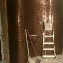 Brązowa mozaika na zaokrąglonej ścianie w trakcie montażu, widoczna drabina, wiadro, szpachelka i przewody na podłodze.