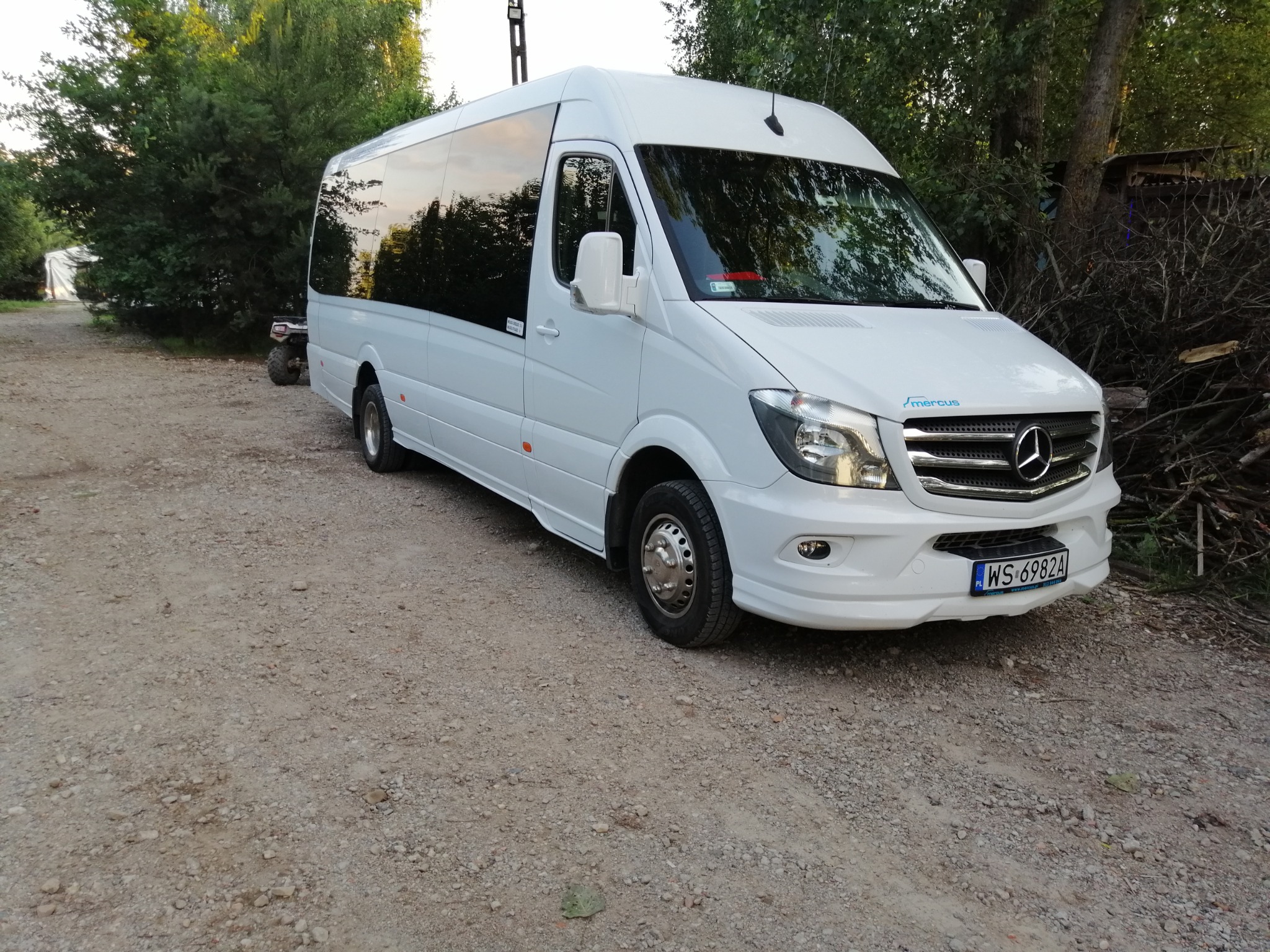 Biały bus marki Mercedes z ciemnymi szybami, zaparkowany na żwirowej drodze, widoczne drzewa w tle i tablica rejestracyjna WS 6982A.