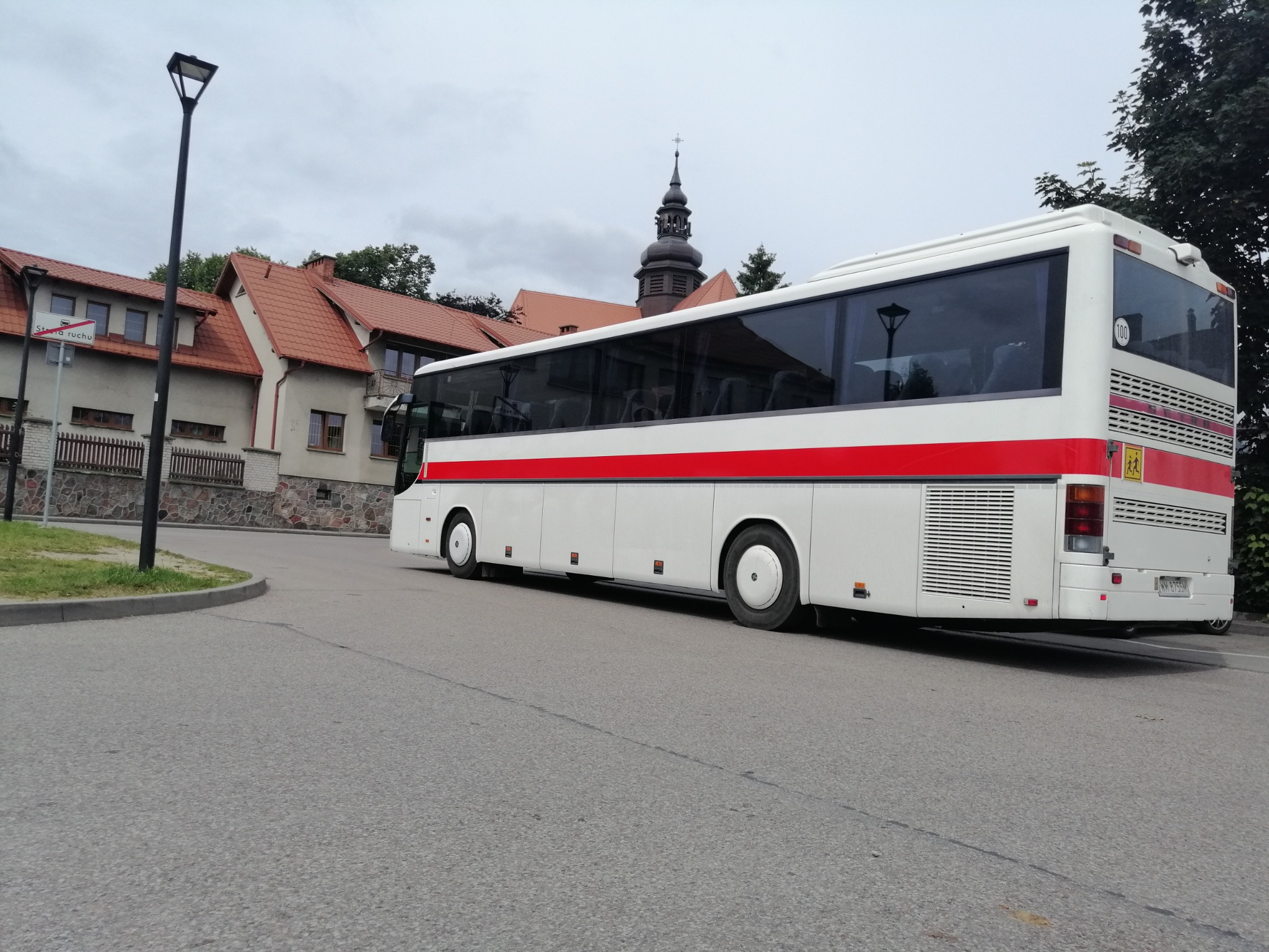 Biały autobus z czerwonym pasem na tle budynku z wieżą kościelną, widziany z perspektywy ulicy.