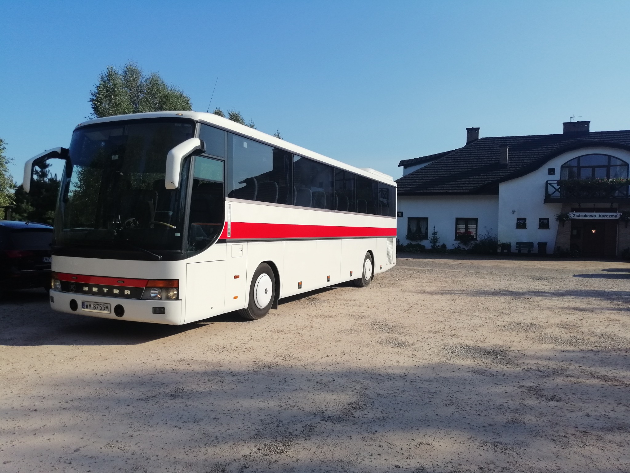 Biały autokar Setra z czerwoną linią na boku zaparkowany na żwirowym placu przed budynkiem z napisem 'Żubiakowa Karczma'.
