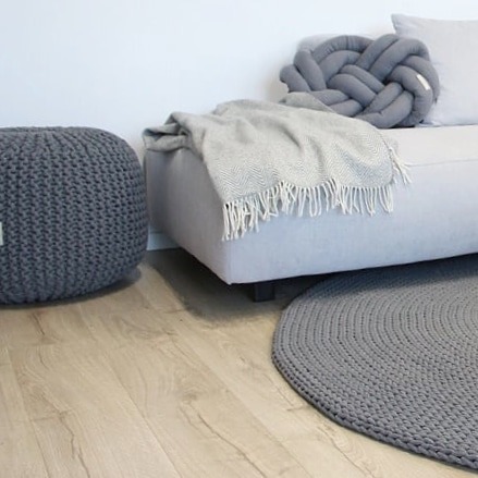 Szary, ręcznie robiony dywan ze sznurka, okrągły, leżący na jasnej podłodze, obok pufa ze sznurka i sofy z poduszką w kształcie węzła.