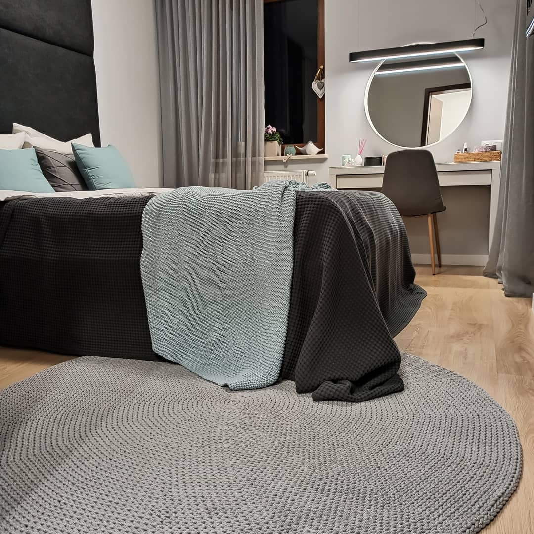 Sypialnia w stonowanej kolorystyce z okrągłym, szarym dywanem ze sznurka, firanami w odcieniach szarości i łóżkiem z turkusowymi poduszkami, oświetlona lampą nad toaletką z okrągłym lustrem.