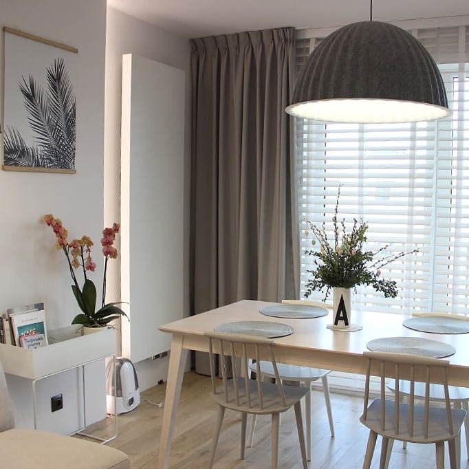 Jasne wnętrze jadalni z drewnianymi żaluzjami w oknach, szarymi zasłonami, białym stołem i krzesłami oraz lampą wiszącą w kształcie kopuły.