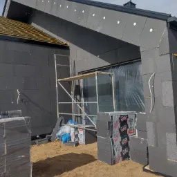 Dom w trakcie ocieplania grafitowym styropianem, widoczne rusztowanie, zabezpieczone okno folią, paczki styropianu na piasku.
