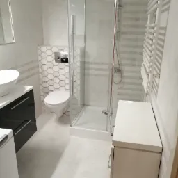 Nowoczesna łazienka w odcieniach bieli i szarości z kabiną prysznicową, podwieszaną toaletą i umywalką nablatową na szafce z czarnymi szufladami, grzejnik drabinkowy.