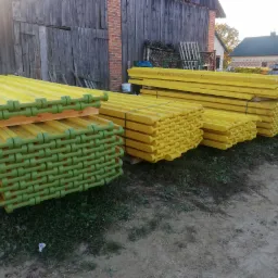 Stosy żółtych, nowych dźwigarów szalunkowych z zielonymi zakończeniami, leżące na ziemi przed drewnianym budynkiem gospodarczym i domem w tle.