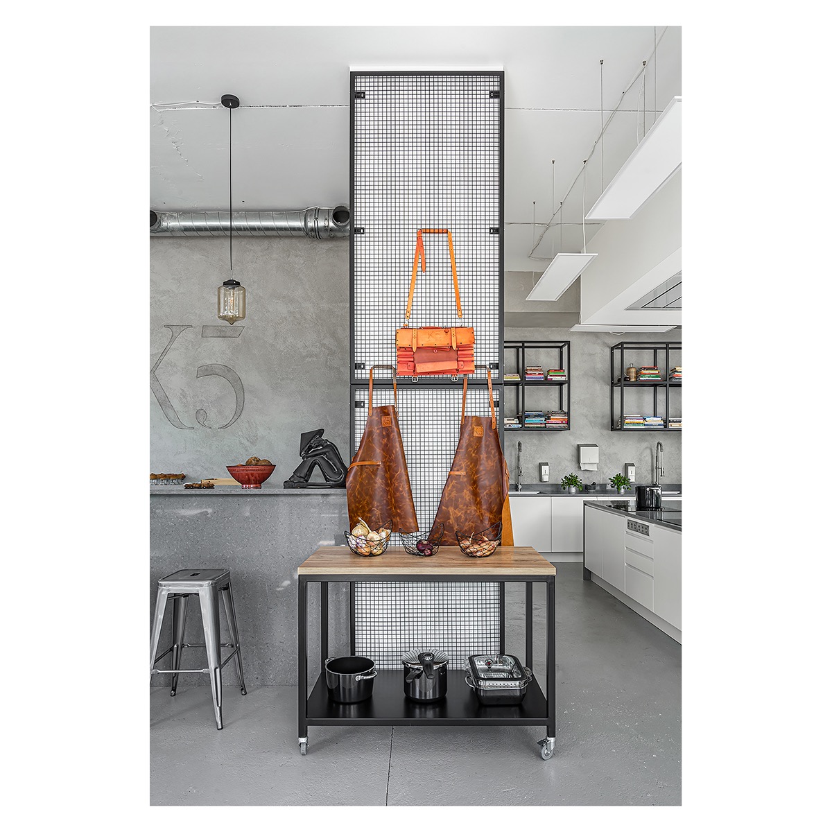 Nowoczesna kuchnia w stylu loft: betonowa ściana z numerem K5, metalowa kratka z wiszącymi skórzanymi fartuchami i torebką, wyspa kuchenna na kółkach z garnkami, stalowy stołek barowy.
