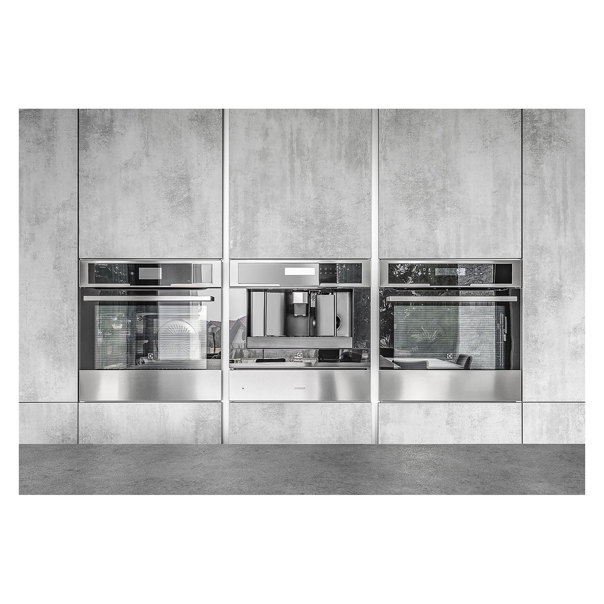 Wbudowane urządzenia kuchenne: piekarniki i ekspres do kawy w obudowie z szarego betonu architektonicznego, z elementami ze stali nierdzewnej.