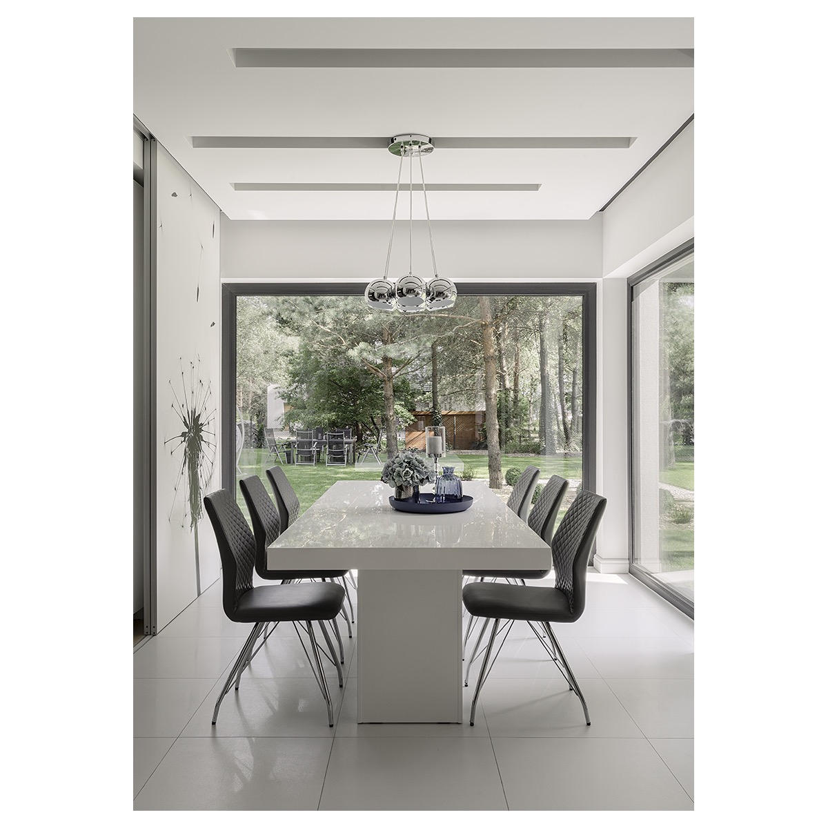Jasna jadalnia z białym stołem, szarymi krzesłami i widokiem na ogród przez duże okno, oświetlona wiszącą lampą z kulistymi kloszami.