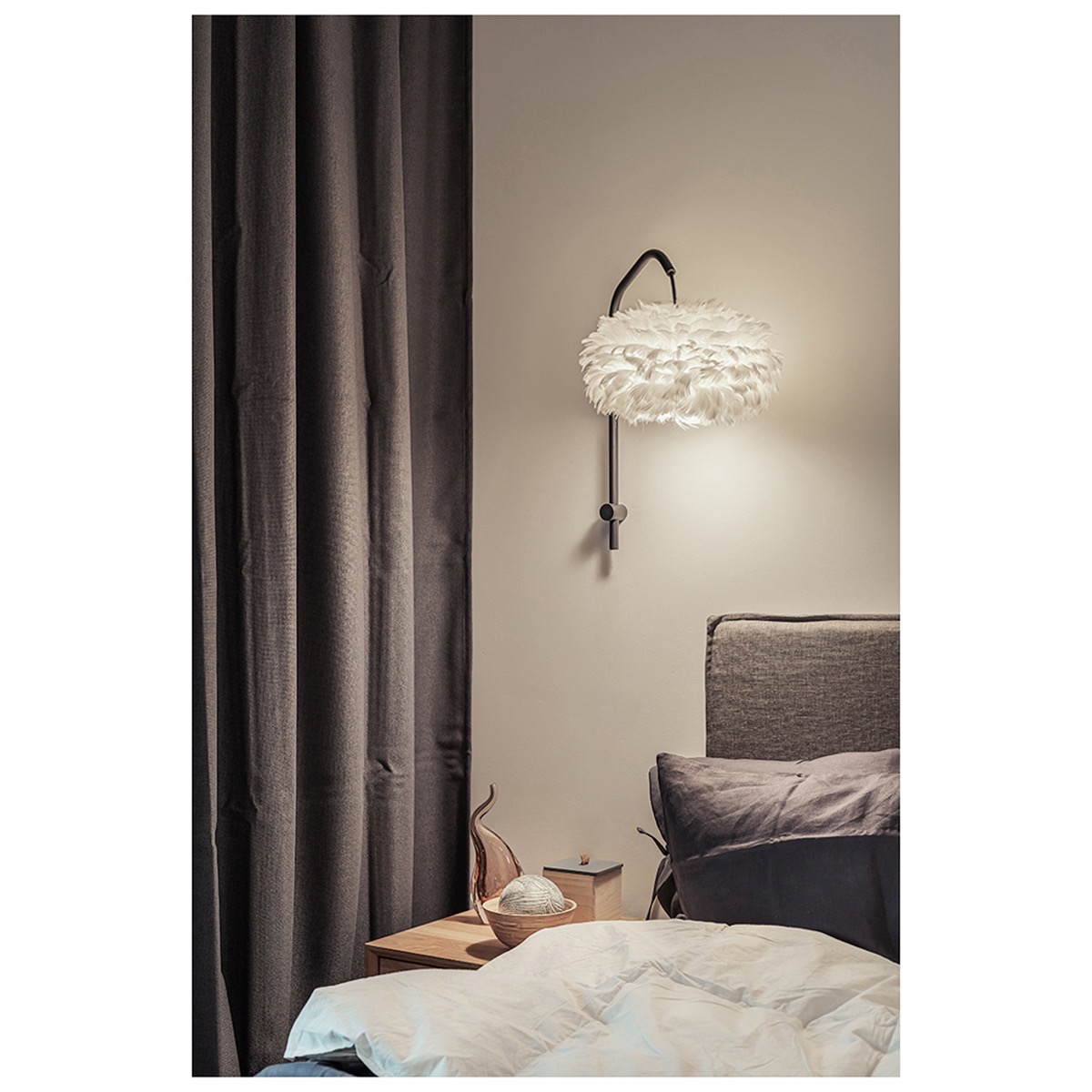 Sypialnia z lampą ścienną w stylu pióra, zasłony w kolorze grafitowym, drewniany stolik nocny z dekoracjami i pościel w odcieniach bieli i szarości.