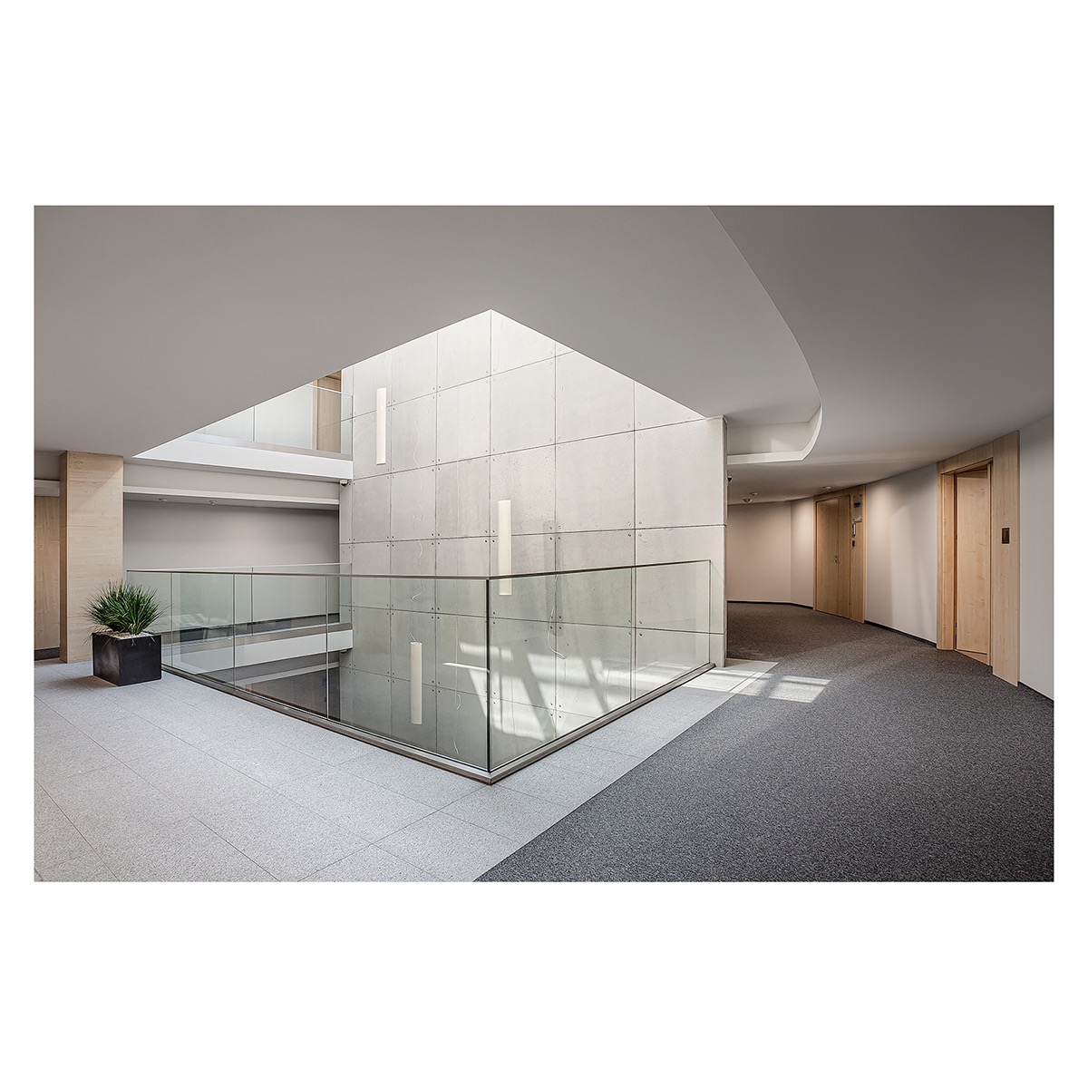 Wnętrze nowoczesnego biurowca z atrium: szara wykładzina, jasne płytki podłogowe, przeszklona balustrada, betonowa ściana z pionowymi panelami oświetleniowymi i donica z rośliną.