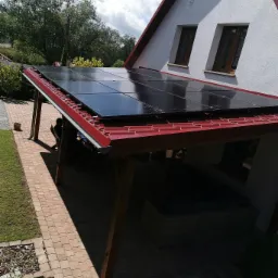 Zadaszenie tarasu z panelami fotowoltaicznymi, widok na dom z czerwonym dachem i dwa okna, kostka brukowa na podłożu.