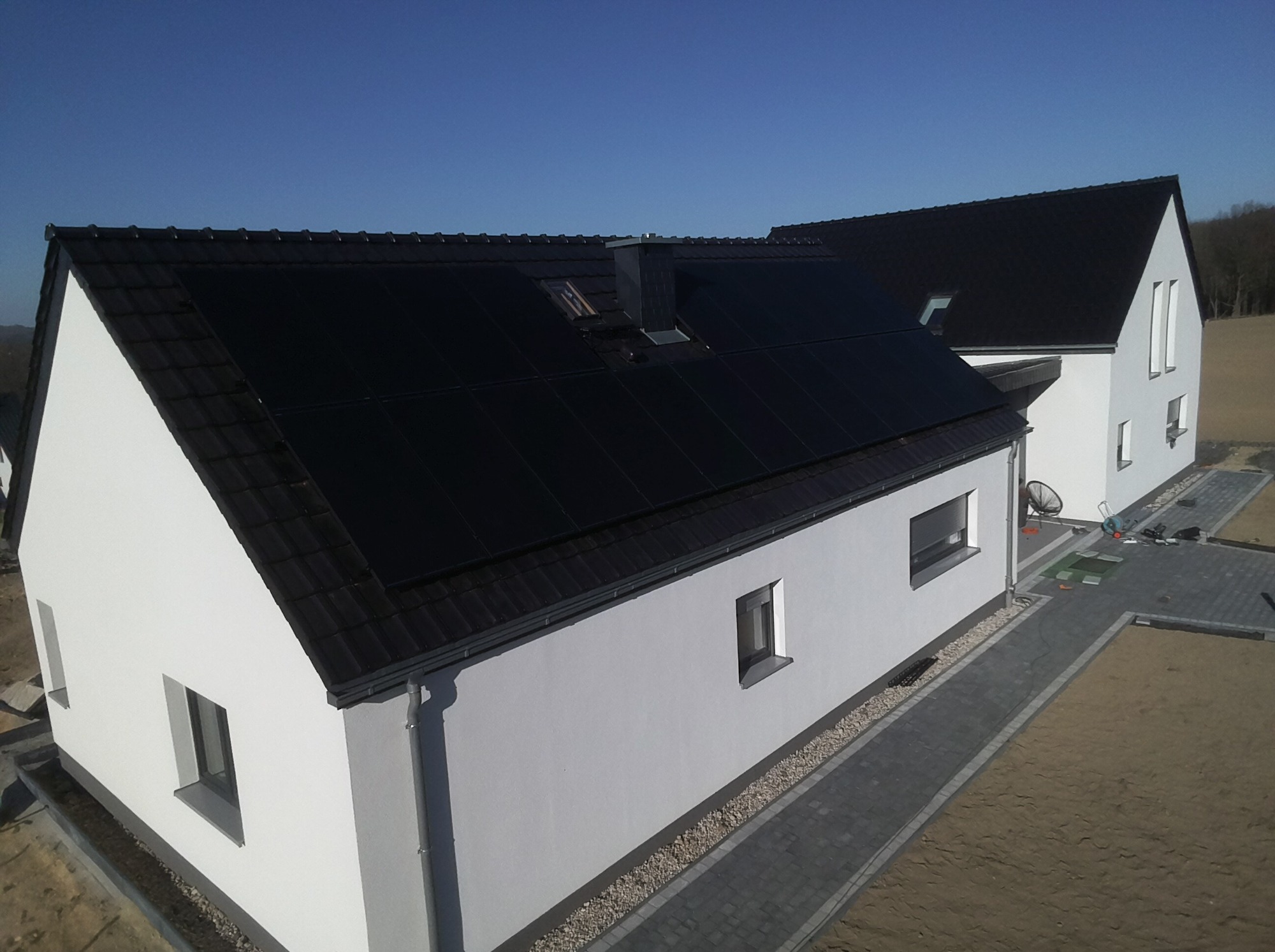 Nowoczesny dom jednorodzinny z białymi ścianami i ciemną dachówką, na której zamontowane są panele słoneczne, widok z góry na ukończoną instalację.