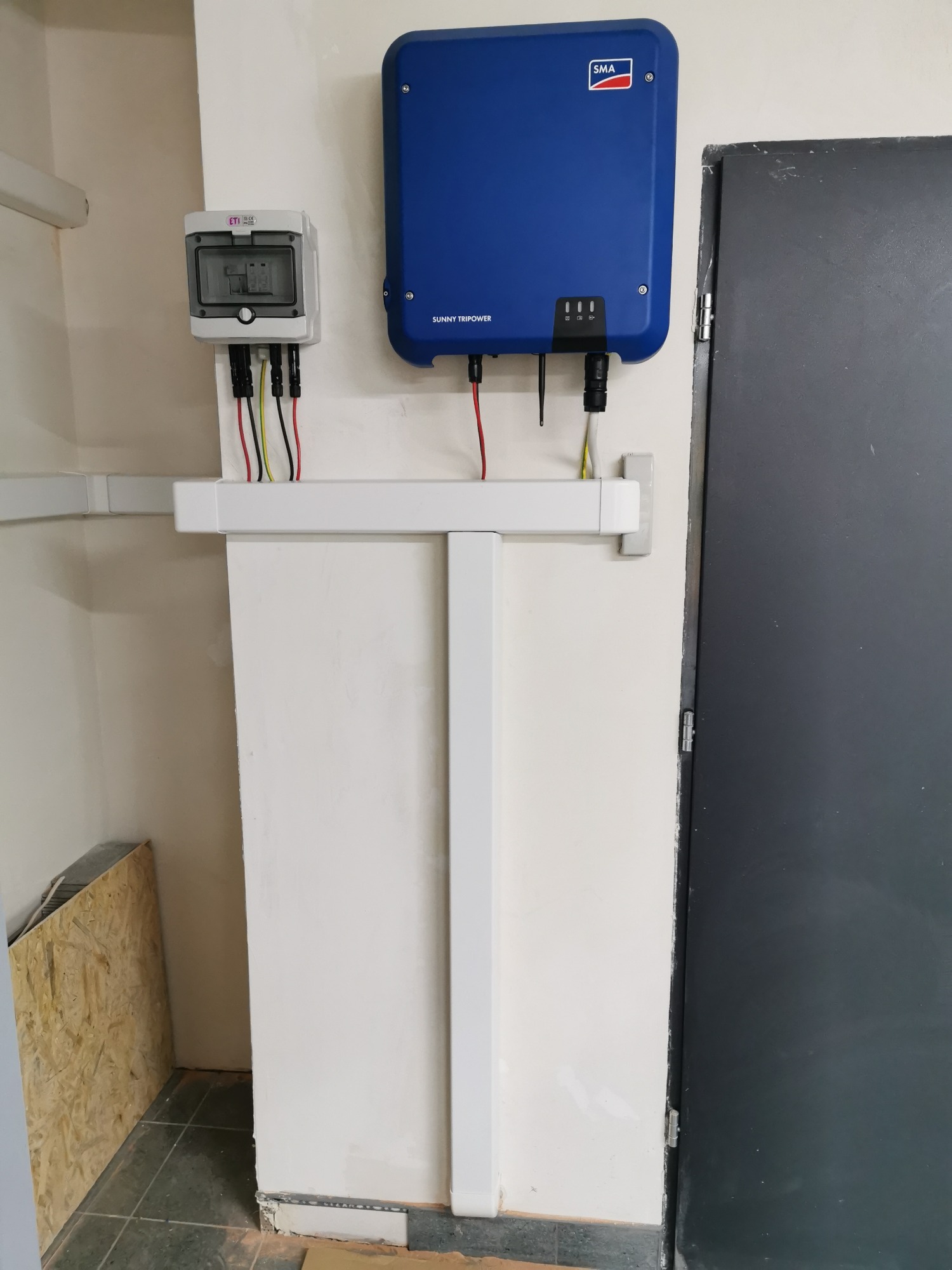 Niebieski falownik SMA Sunny Tripower zamontowany na białej ścianie, obok szara skrzynka z bezpiecznikami ETI i czarnymi przewodami, białe korytko kablowe, widoczny fragment ściany z surowym...