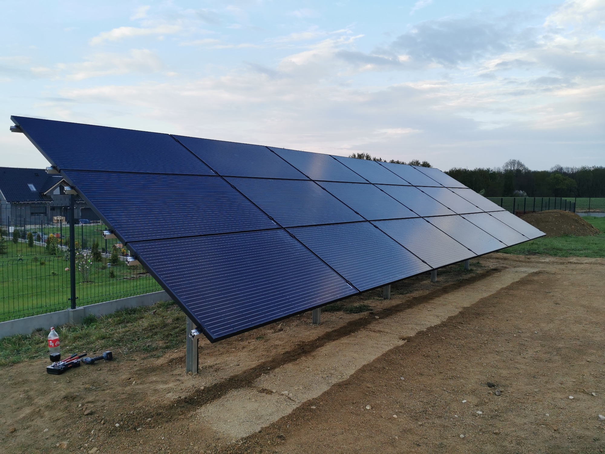 Panel słoneczny na metalowych wspornikach, częściowo zamontowany na placu budowy; widoczne narzędzia i butelka napoju, w tle dom i siatka ogrodzeniowa.