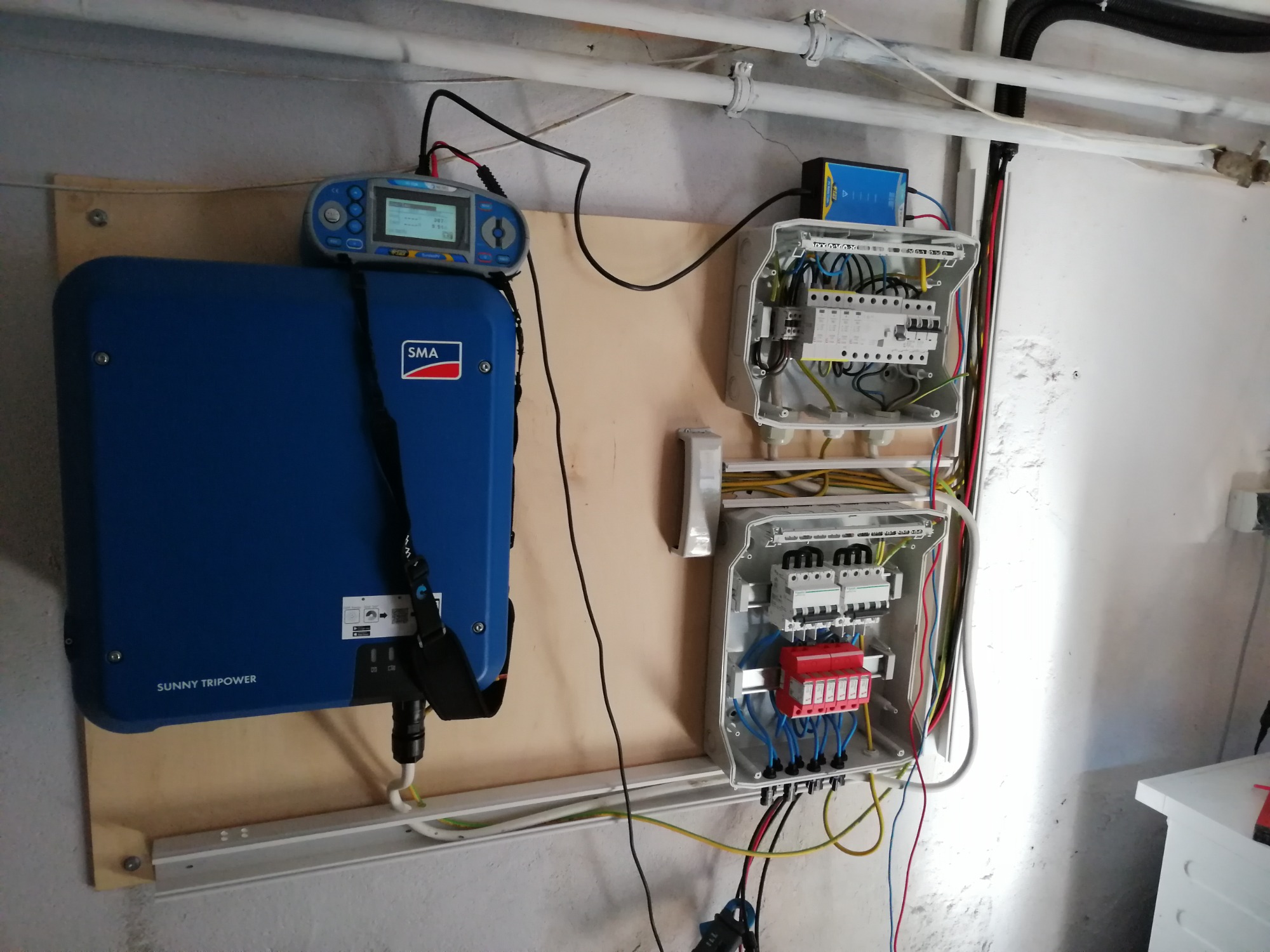 Niebieski falownik SMA Sunny Tripower zamontowany na sklejce obok dwóch otwartych skrzynek elektrycznych z przewodami i zabezpieczeniami, dodatkowo widoczny miernik elektryczny.