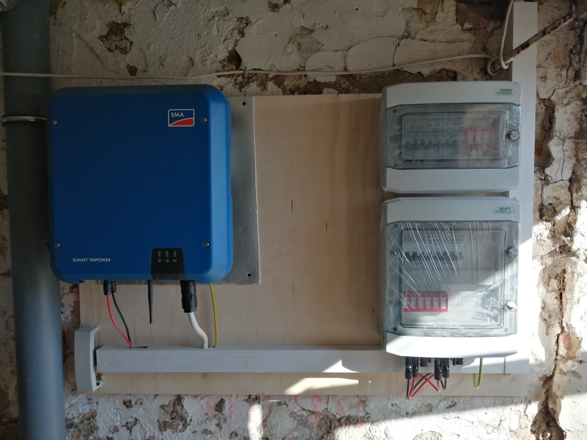Niebieski falownik SMA Sunny Tripower zamontowany obok dwóch rozdzielnic elektrycznych NOARK w plastikowej folii ochronnej, na drewnianej płycie przytwierdzonej do ściany z odpadającym tynkiem.