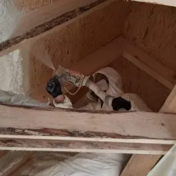 Pracownik w kombinezonie ochronnym aplikuje piankę izolacyjną w przestrzeni poddasza, widoczne drewniane krokwie i warstwa pianki na folii.