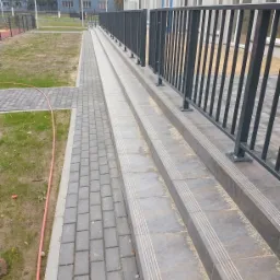 Zewnętrzne schody z szarej kostki brukowej i metalową barierką, prowadzące do budynku z niebieskimi i białymi elementami.