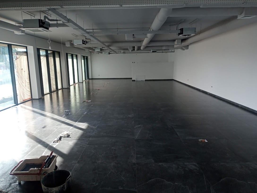 Układanie ciemnych płytek podłogowych w dużym, jasnym pomieszczeniu biurowym z widocznymi narzędziami glazurniczymi i pozostałościami po pracy.