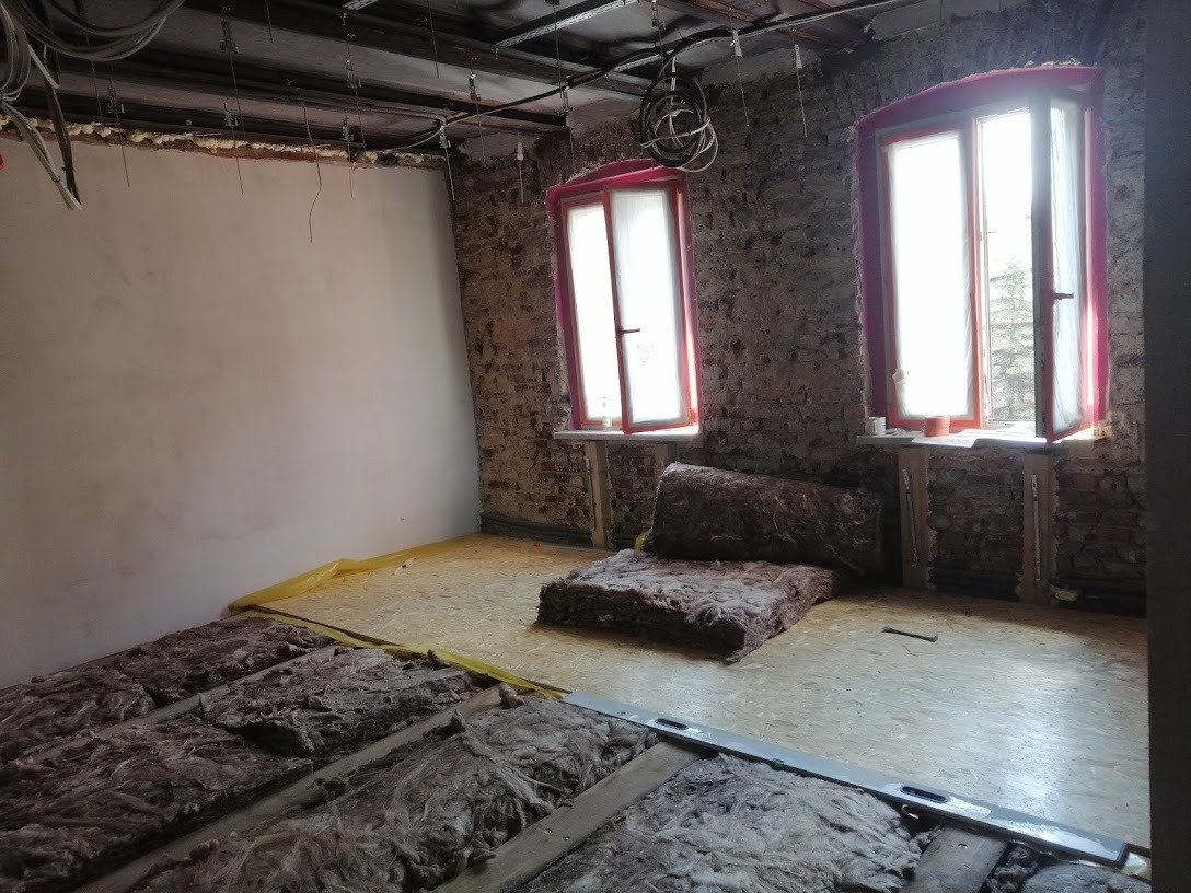 Wnętrze w trakcie remontu: odsłonięta cegła na ścianach, okna z czerwonymi ramami, izolacja z wełny mineralnej na podłodze, widoczne instalacje pod sufitem.
