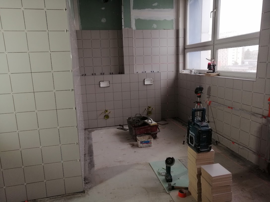 Układanie jasnoszarych płytek w łazience w trakcie remontu, widoczny laser krzyżowy i zapas płytek na podwyższeniu.